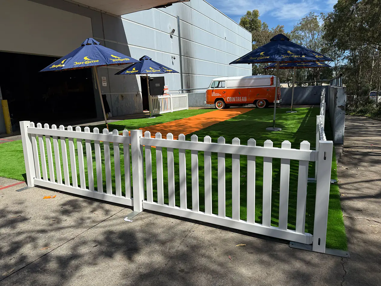 White picket fence panels enclosing an outdoor event display area with artificial grass, branded umbrellas, and a vintage van setup