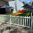 White picket fence panels enclosing an outdoor event display area with artificial grass, branded umbrellas, and a vintage van setup