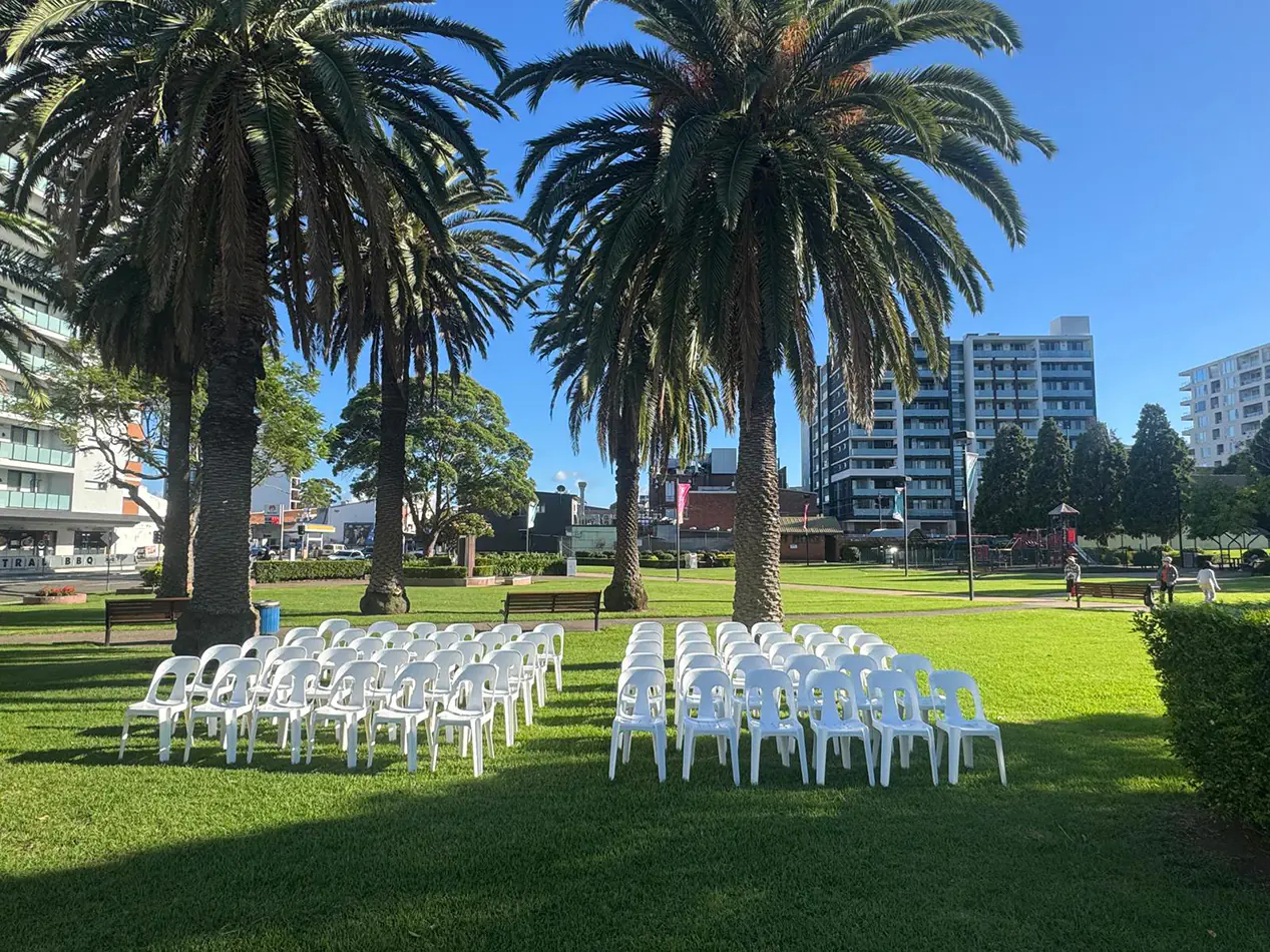 White plastic chairs arranged in rows on grass in a park setting with palm trees and surrounding buildings for an outdoor event