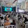 A large crowd of people sitting in white plastic stackable chairs in an auditorium.