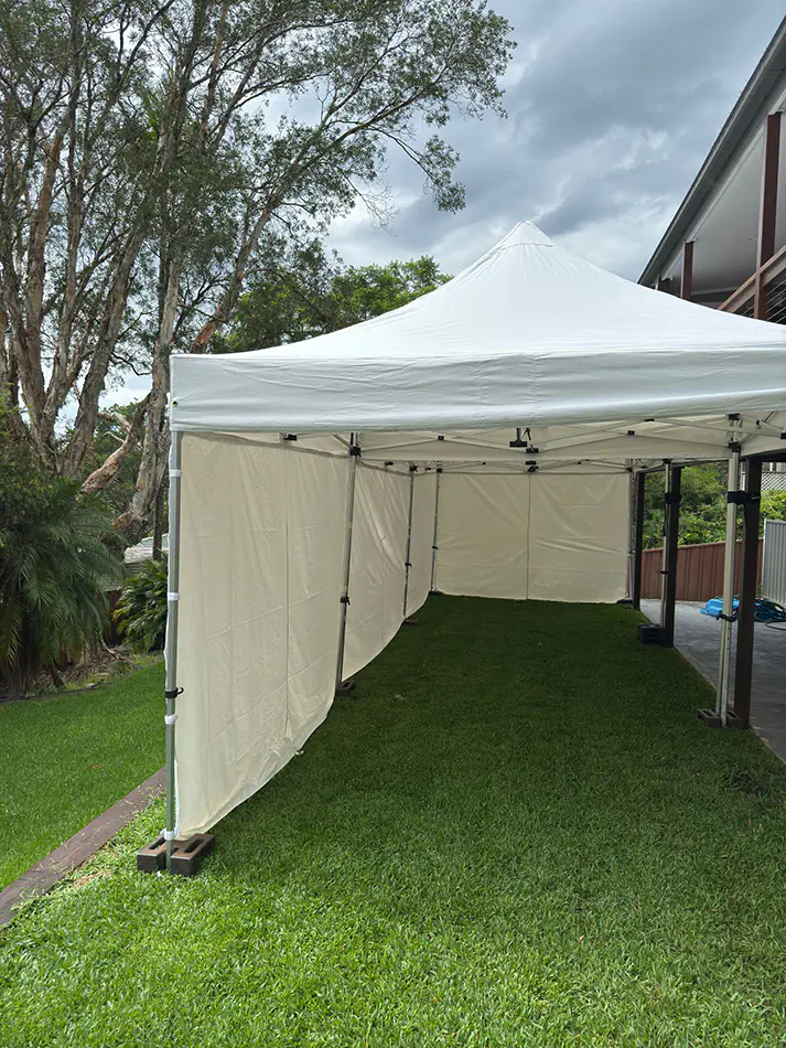 White pop up marquee with side walls set up on grass beside a home, with weighted frame legs, trees and cloudy sky in the background