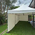 White pop up marquee with side walls set up on grass beside a home, with weighted frame legs, trees and cloudy sky in the background