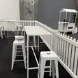 A room adorned with a white picket fence and white stools surrounding a White Rectangular Tapas Table Hire with a white top.