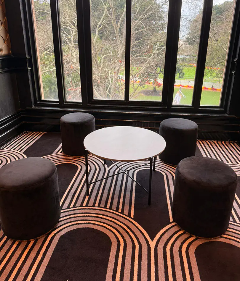 White round cafÃ© table surrounded by black ottoman seating inside a modern lounge space with patterned carpet flooring