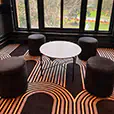 White round cafÃ© table surrounded by black ottoman seating inside a modern lounge space with patterned carpet flooring