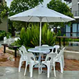 White round café table with plastic chairs under a large outdoor umbrella in a landscaped courtyard setting with greenery and paved flooring