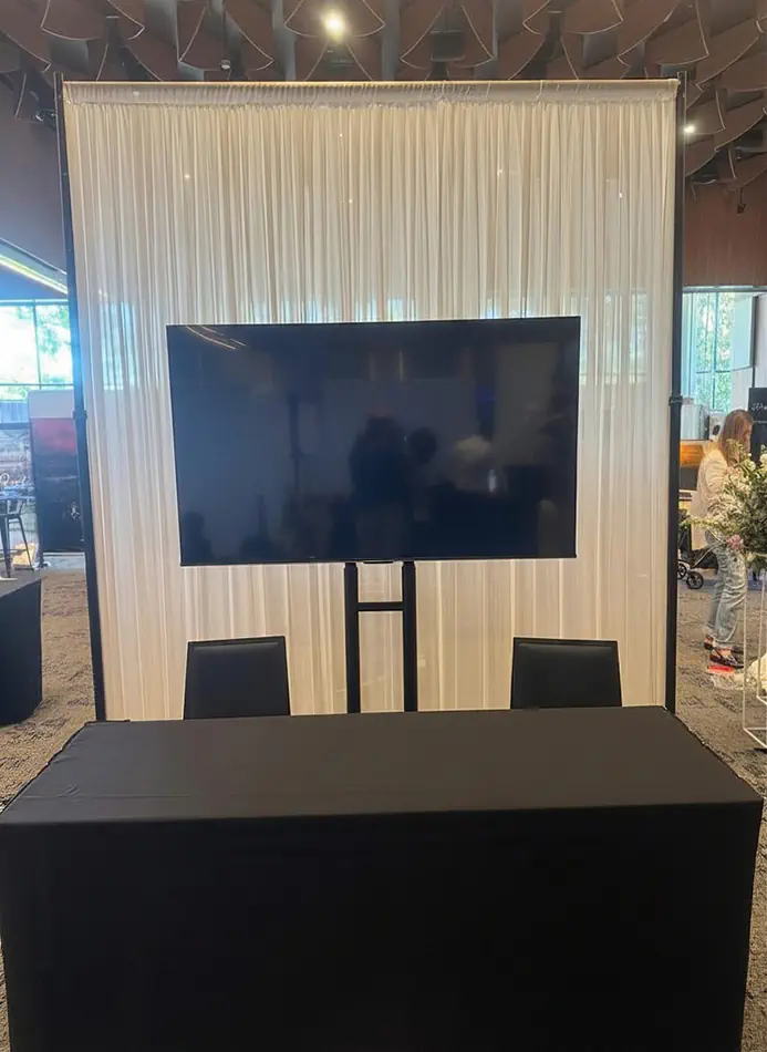 White sheer draping on a frame with a mounted TV screen and black tablecloth display table for an event booth or presentation area