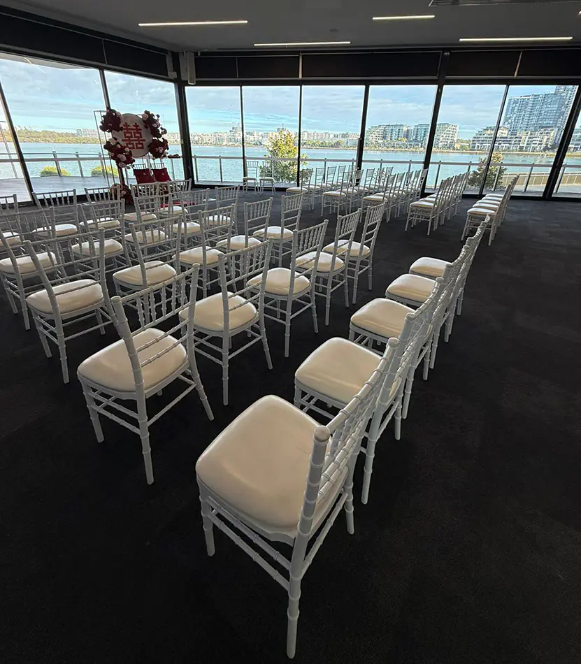 White Tiffany chairs arranged for a waterfront wedding ceremony inside a modern function venue overlooking the water