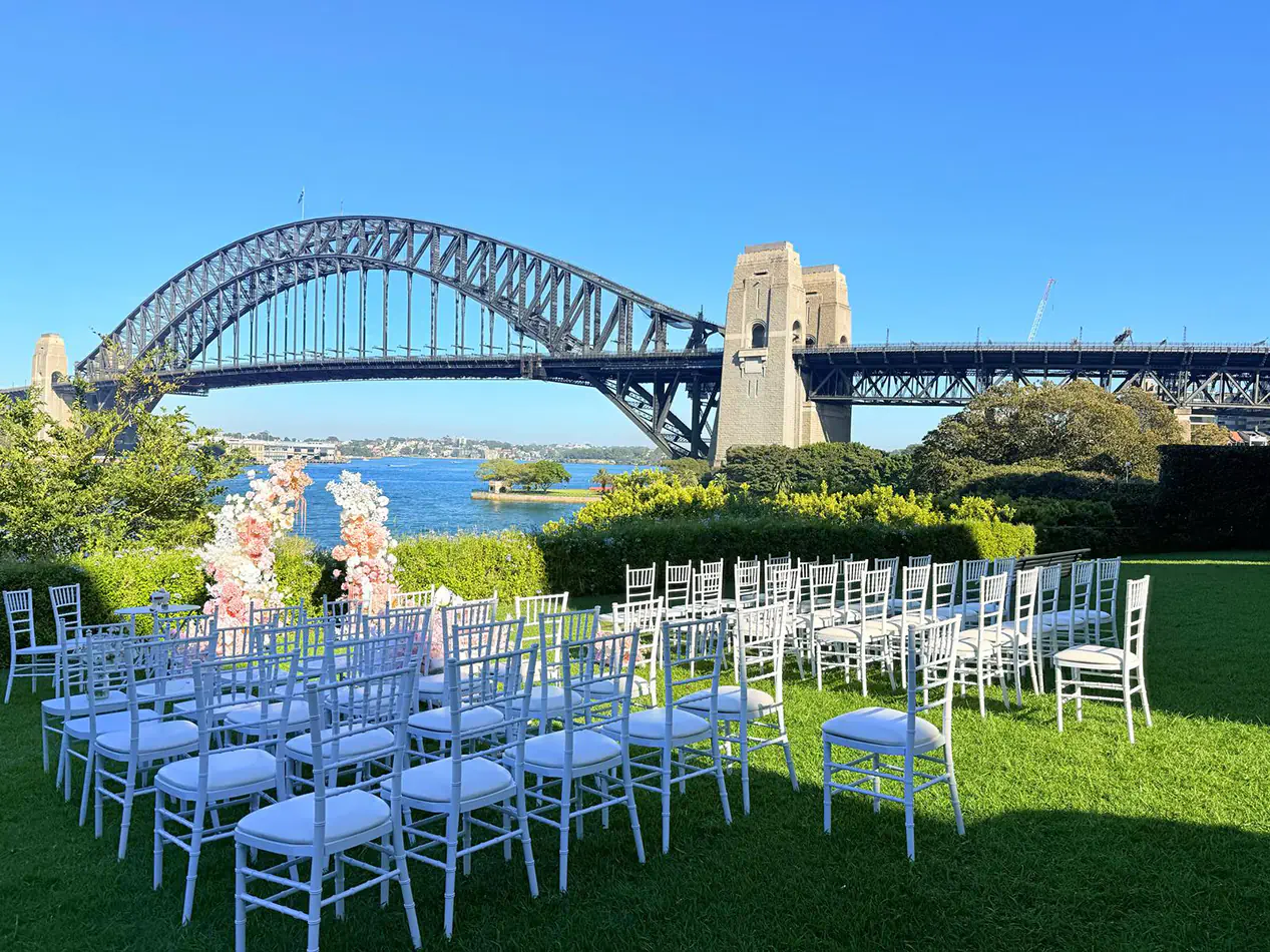 White Tiffany chairs arranged for an outdoor wedding ceremony with Sydney Harbour Bridge and waterfront views in the background