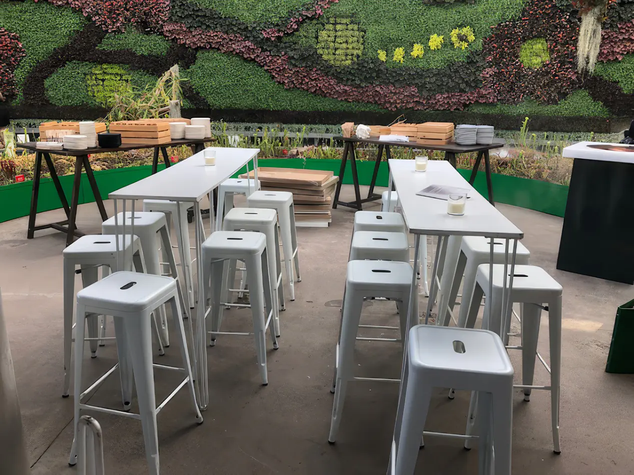 White Tolix stools arranged around long white bar tables in an outdoor garden event space with a green vertical wall backdrop