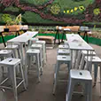 White Tolix stools arranged around long white bar tables in an outdoor garden event space with a green vertical wall backdrop
