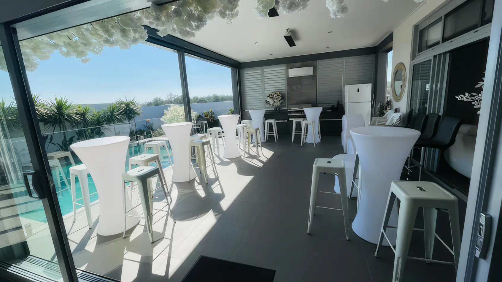 White Tolix stools paired with white cocktail tables on a bright covered balcony event area overlooking a swimming pool and outdoor view