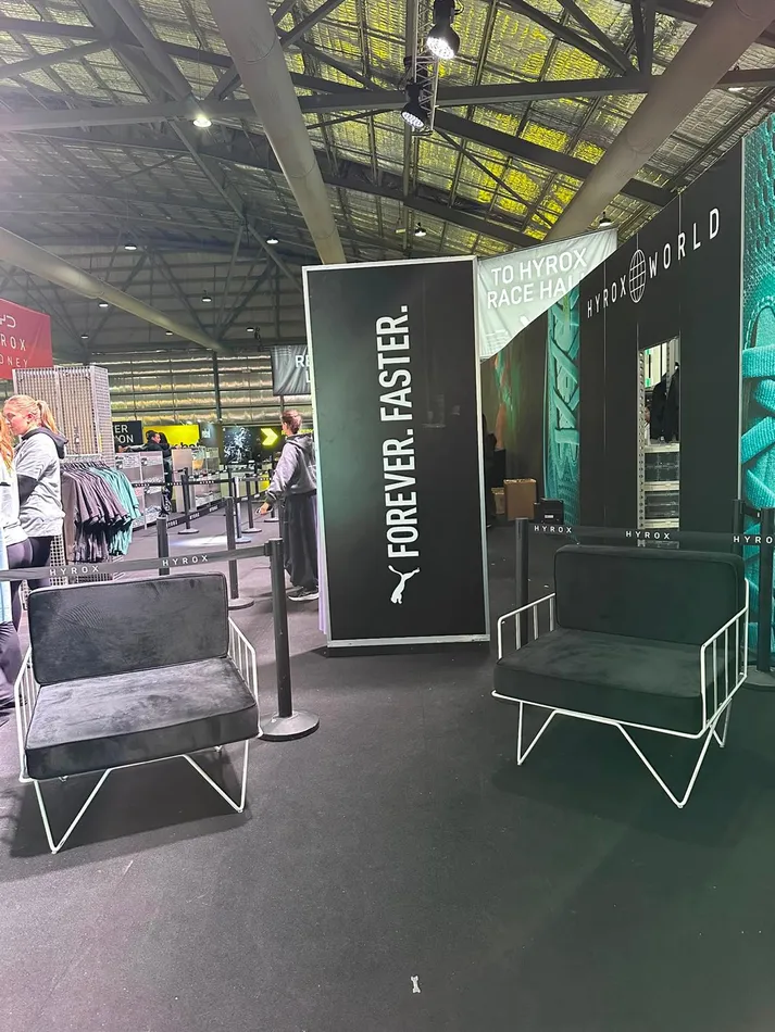 Two white wire arm chairs with black velvet cushions set up as a lounge area at an indoor fitness expo