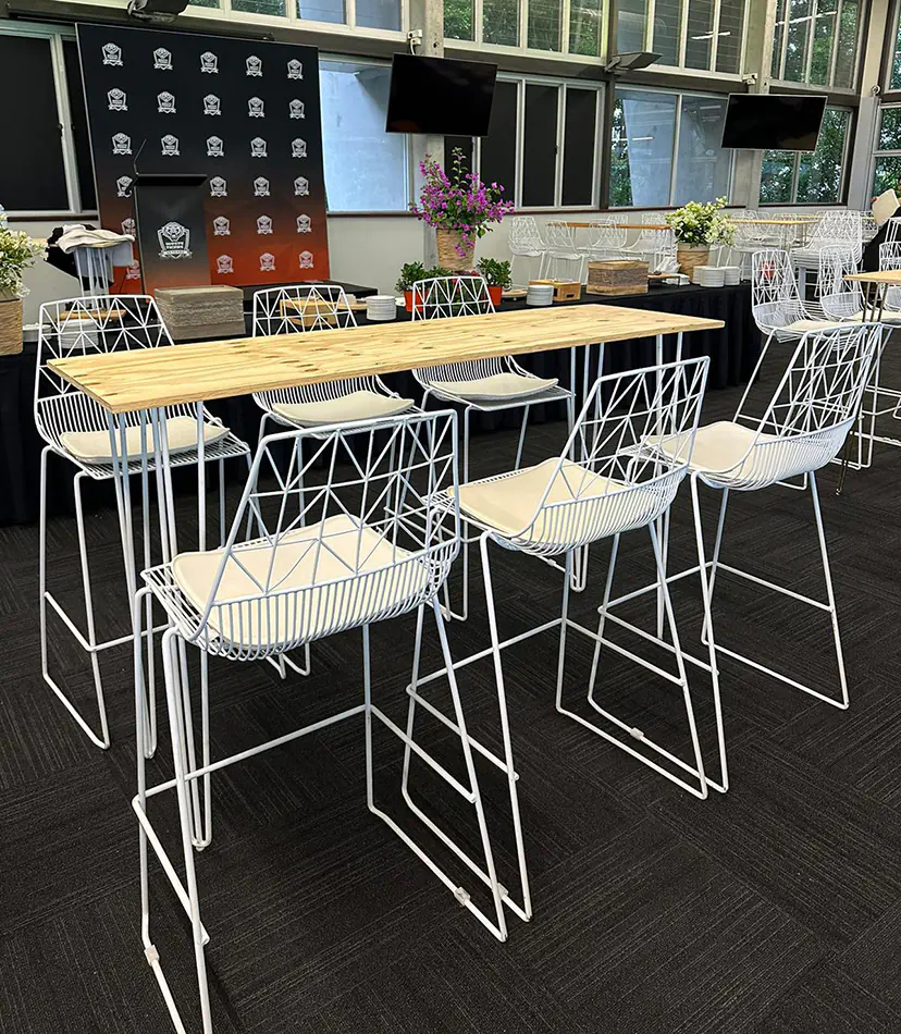 White wire bar chairs arranged around a long timber top bar table in an indoor event space with buffet setup, presentation backdrop, and large windows