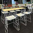 White wire bar chairs arranged around a long timber top bar table in an indoor event space with buffet setup, presentation backdrop, and large windows