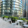 A rooftop patio with chairs and white wire cocktail tables in front of tall buildings.
