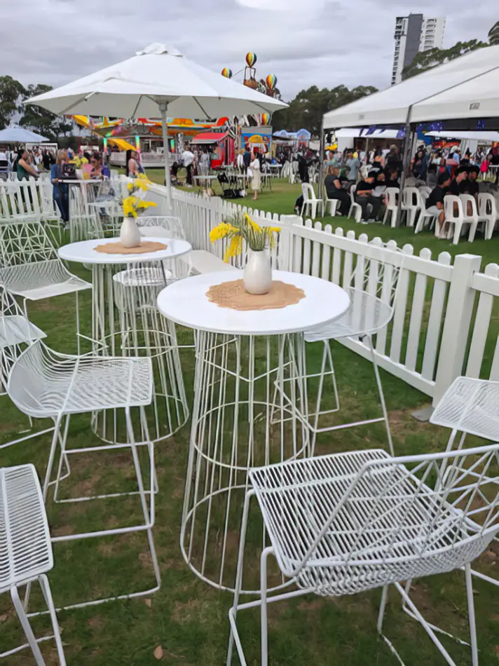 White wire cocktail table hire with matching stools arranged at an outdoor festival event, featuring umbrellas, fencing and crowd seating areas