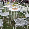 White wire cocktail table hire with matching stools arranged at an outdoor festival event, featuring umbrellas, fencing and crowd seating areas