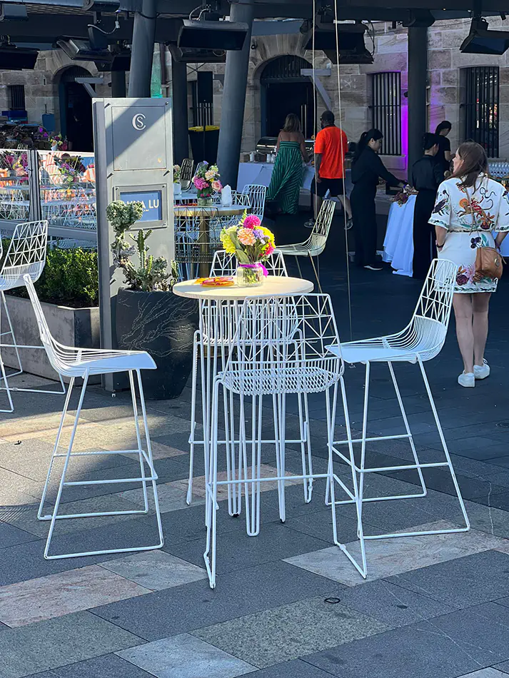 White wire cocktail table with matching bar stools set up outdoors at an event space with floral centrepiece and casual bar seating arrangement