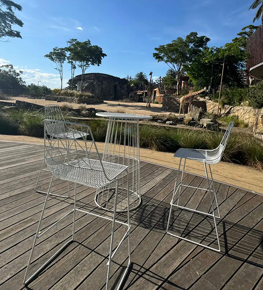 White wire cocktail table with matching white wire bar stools on an outdoor timber deck overlooking landscaped gardens and animal enclosure displays