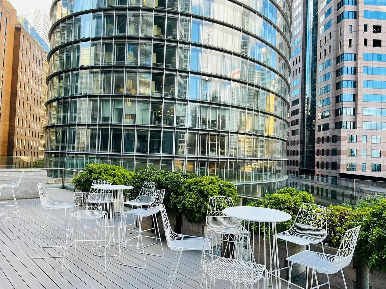 A rooftop patio with chairs and white wire cocktail tables in front of tall buildings.
