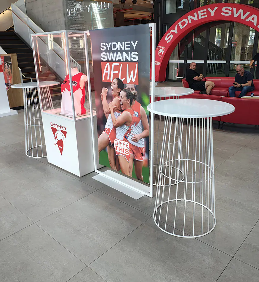 White wire cocktail tables displayed inside the Sydney Swans AFLW event space with branded promotional displays and lounge seating