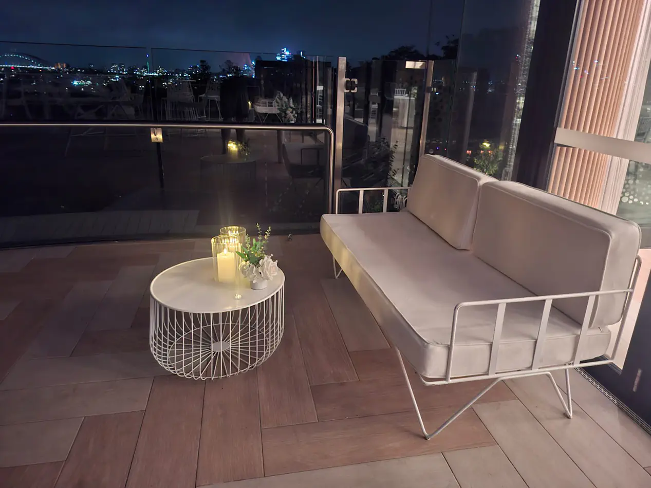 White wire coffee table with candle styling and white lounge seating arranged on a balcony event space overlooking the Sydney skyline at night