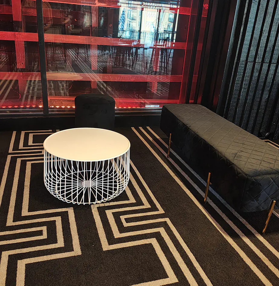White wire coffee table paired with black velvet ottoman seating inside a modern event lounge with geometric patterned carpet and red lighting