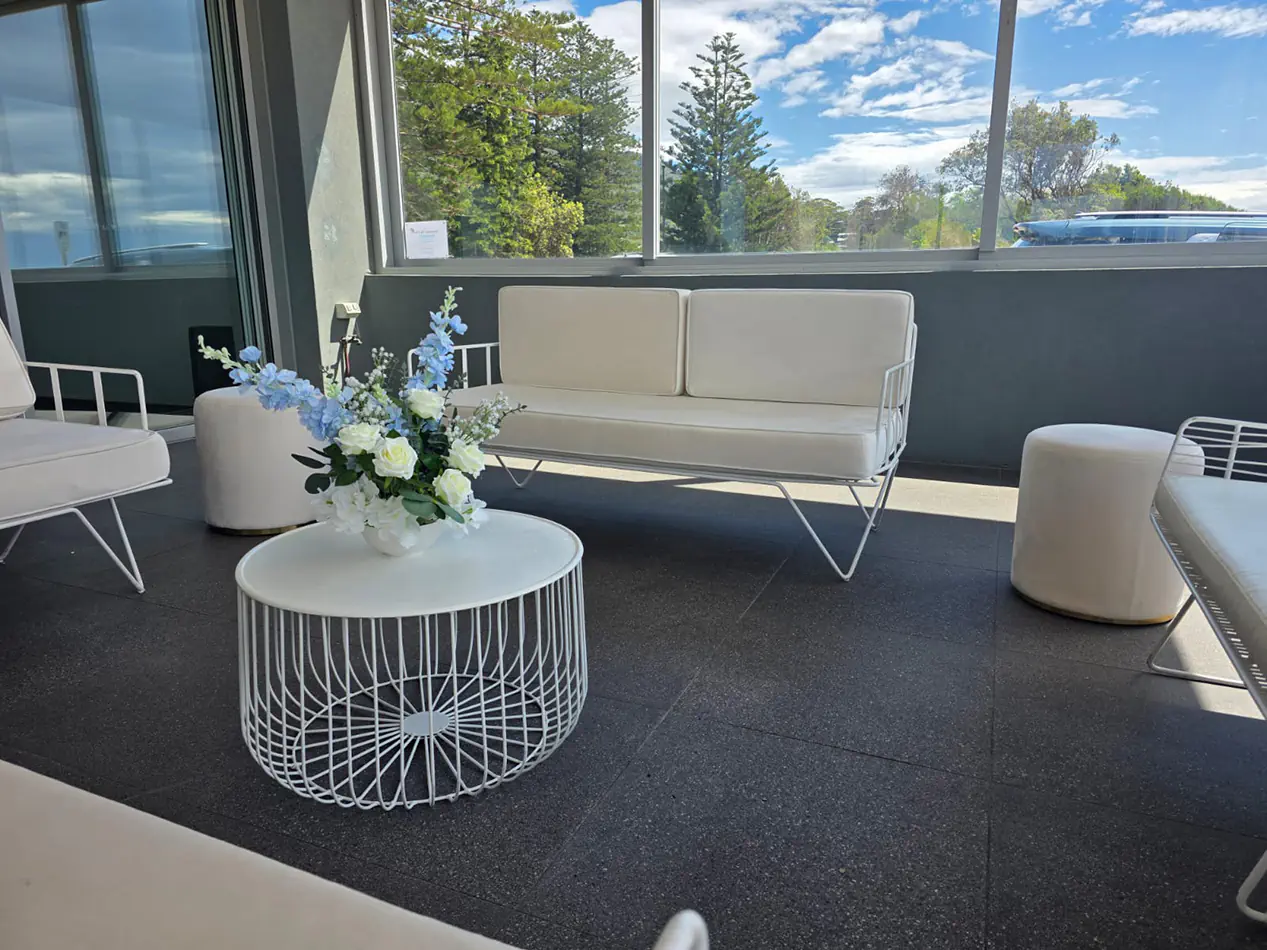 White wire coffee table with white lounge seating and velvet ottoman stools styled inside a bright modern event venue with floral centrepiece