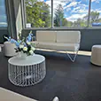 White wire coffee table with white lounge seating and velvet ottoman stools styled inside a bright modern event venue with floral centrepiece