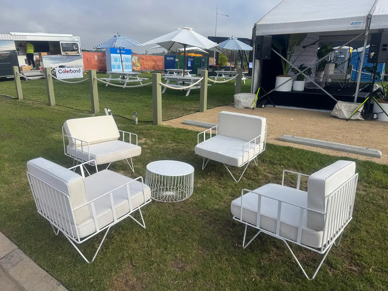 White wire lounge chairs with matching cushions and round wire coffee table styled on grass beside an outdoor marquee event setup