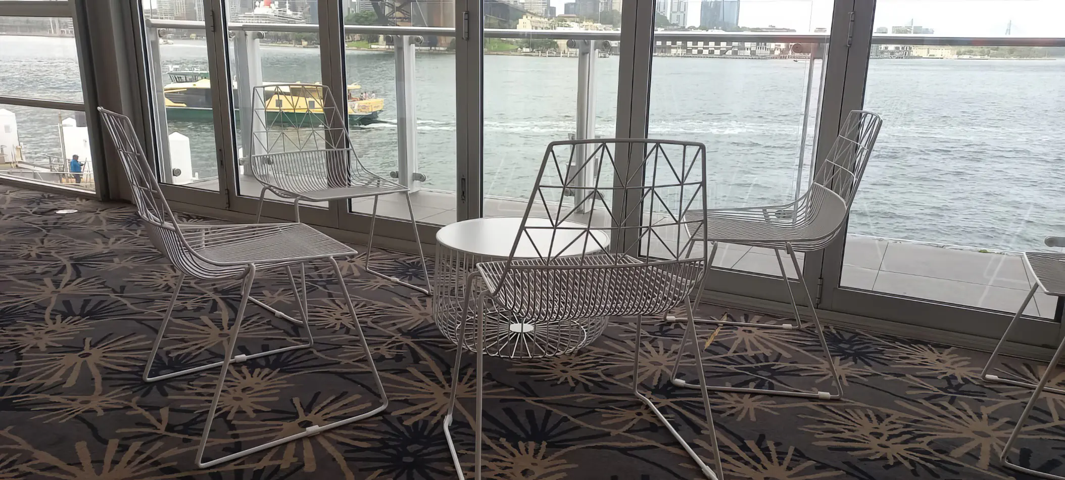 White wire lounge chairs and matching round coffee table positioned indoors beside large windows overlooking a waterfront harbour view