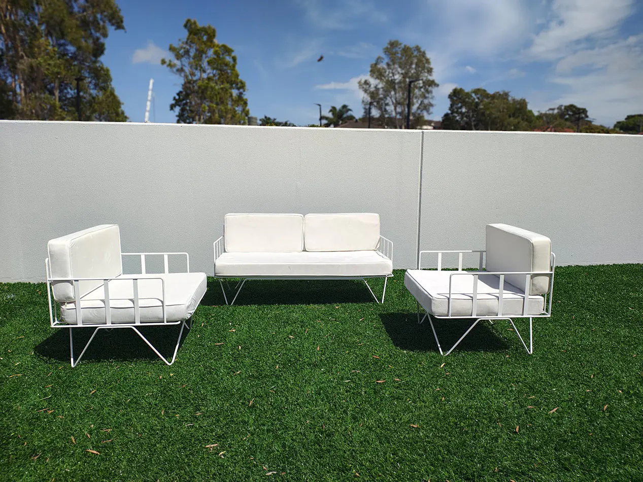White wire lounge sofa and matching armchairs with white cushions arranged on artificial grass in an outdoor event setting with modern white wall backdrop