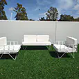 White wire lounge sofa and matching armchairs with white cushions arranged on artificial grass in an outdoor event setting with modern white wall backdrop