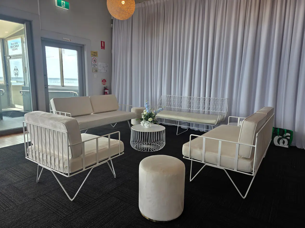 White wire lounge furniture with velvet cushions, round wire coffee table and ottoman stools styled inside a wedding and cocktail reception venue with white draped backdrop