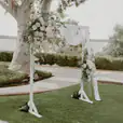 White wooden arbor decorated with soft white and blush floral arrangements and draped fabric, set up on a lawn for an outdoor wedding ceremony with trees and water views in the background.