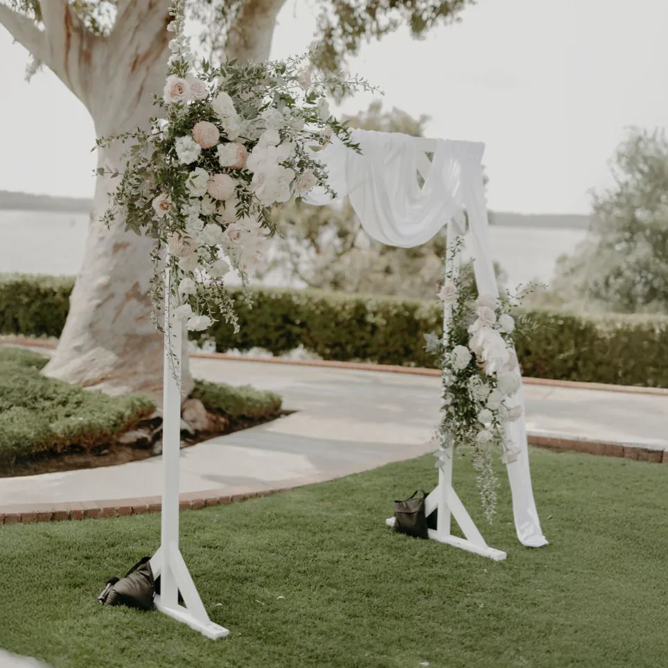 White wooden arbor decorated with soft white and blush floral arrangements and draped fabric, set up on a lawn for an outdoor wedding ceremony with trees and water views in the background.