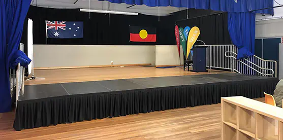 Auditorium stage extension with black skirting, side stairs, safety railings, lectern, flags and blue curtains in a school presentation hall