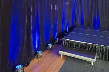 Black stage draping with blue uplighting, timber flooring, black skirted stage platform and front access step in an indoor event room
