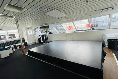 Black stage platform installed inside a boat function room, with low modular staging, wraparound windows and lounge seating nearby