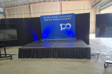 Corporate stage setup with black draping, blue uplighting, branded backdrop display, black skirted platform and side screens inside an industrial event space