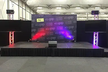 Event stage platform with black skirting, branded backdrop wall, side screens, truss lighting and speakers inside a large indoor venue