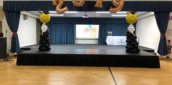 Graduation stage extension with black skirting, projector screen, blue curtains, balloon columns and gold 2019 number balloons in a school hall