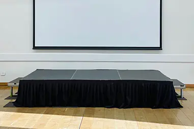 Indoor stage platform positioned in front of a large projector screen for a presentation or school event, with black skirting and timber flooring