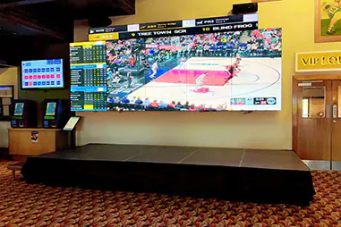 Indoor stage platform set up beneath a large video wall and betting display screens beside a VIP lounge entrance in an entertainment venue