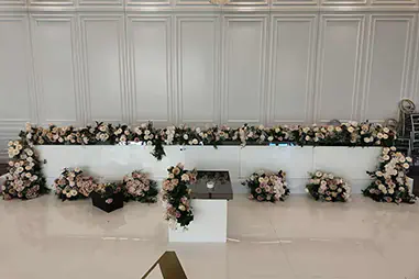 Luxury wedding stage with a long white bridal table, blush floral arrangements, glossy white flooring, chandeliers and detailed wall panelling in a formal reception venue