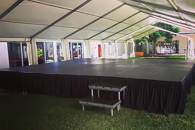 Marquee covered stage platform with black skirting, access step and large sheltered performance area set up on grass beside a building