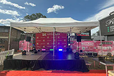 Outdoor marquee stage with black skirting, branded backdrop, speakers, coloured lighting and event equipment set up on a red carpet area