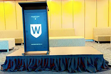 Podium hire setup with a black skirted presentation platform, branded lectern, carpeted flooring and lounge seating in a modern indoor venue