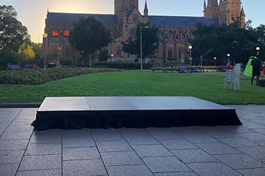 Portable black stage platform set on a paved outdoor area overlooking a lawn and a large cathedral building at dusk